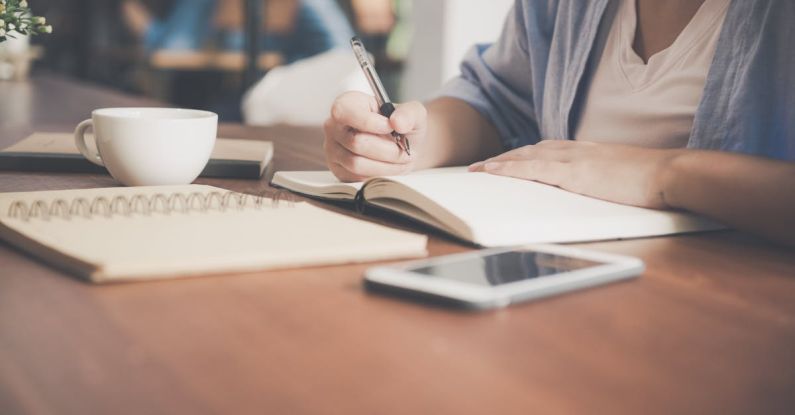 Tech Banking - Woman Writing on a Notebook Beside Teacup and Tablet Computer