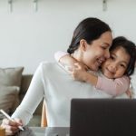 Child Digital - Happy young Asian woman working remotely from home with laptop and tablet while adorable little daughter hugging from behind
