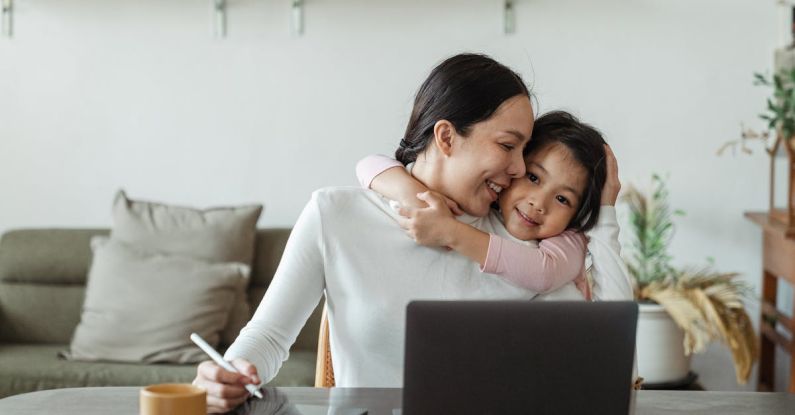 Child Digital - Happy young Asian woman working remotely from home with laptop and tablet while adorable little daughter hugging from behind