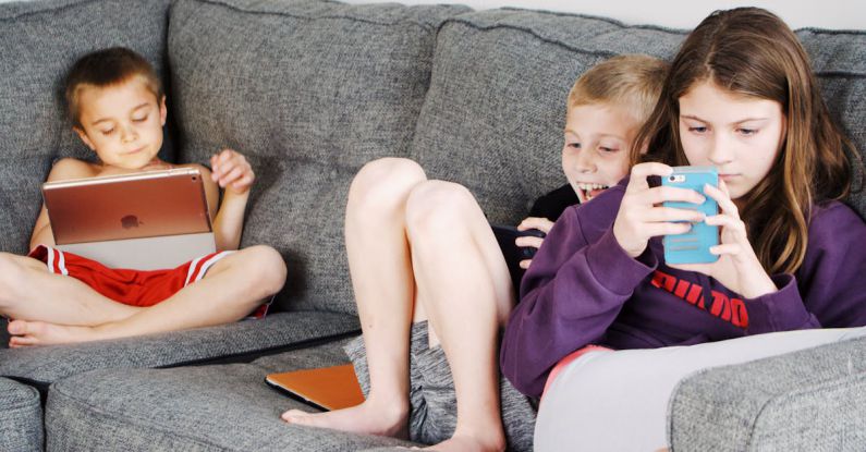 Positive Habits - Positive barefoot children in casual wear resting together on cozy couch and browsing tablets and smartphones