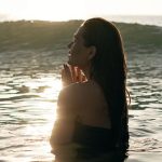 Rest Relaxation - Side view of tranquil young female tourist with long dark hair standing in waving ocean with closed eyes and enjoying summer sunset