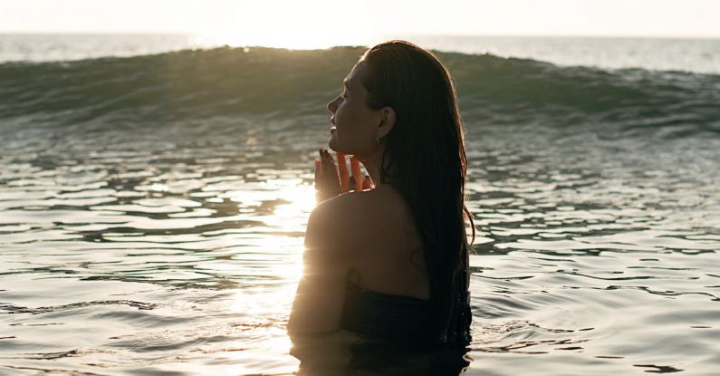 Rest Relaxation - Side view of tranquil young female tourist with long dark hair standing in waving ocean with closed eyes and enjoying summer sunset