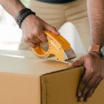 Mortgage Process - Crop faceless young male with wristwatch using adhesive tape while preparing cardboard box for transportation