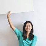 Stocks Real Estate - Cheerful Asian woman sitting cross legged on floor against white wall in empty apartment and showing white blank banner