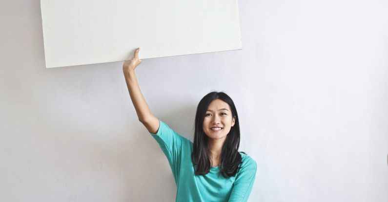 Stocks Real Estate - Cheerful Asian woman sitting cross legged on floor against white wall in empty apartment and showing white blank banner