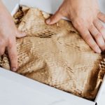 E-commerce Store - From above crop adult male packing ordered product or gift in white cardboard box with craft paper for safety postal delivery at white marble table